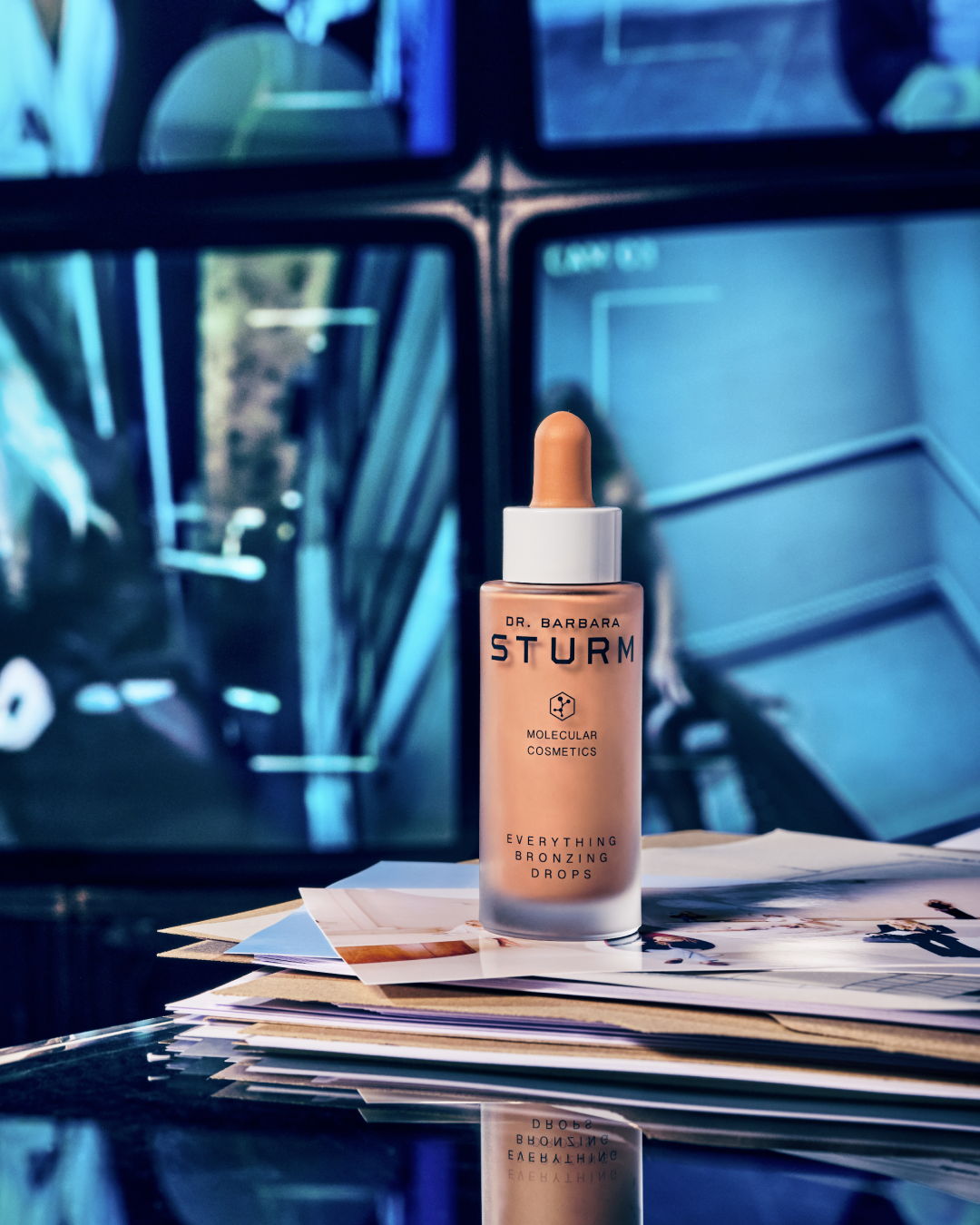 A skincare serum bottle on a table with papers and monitors in the background.