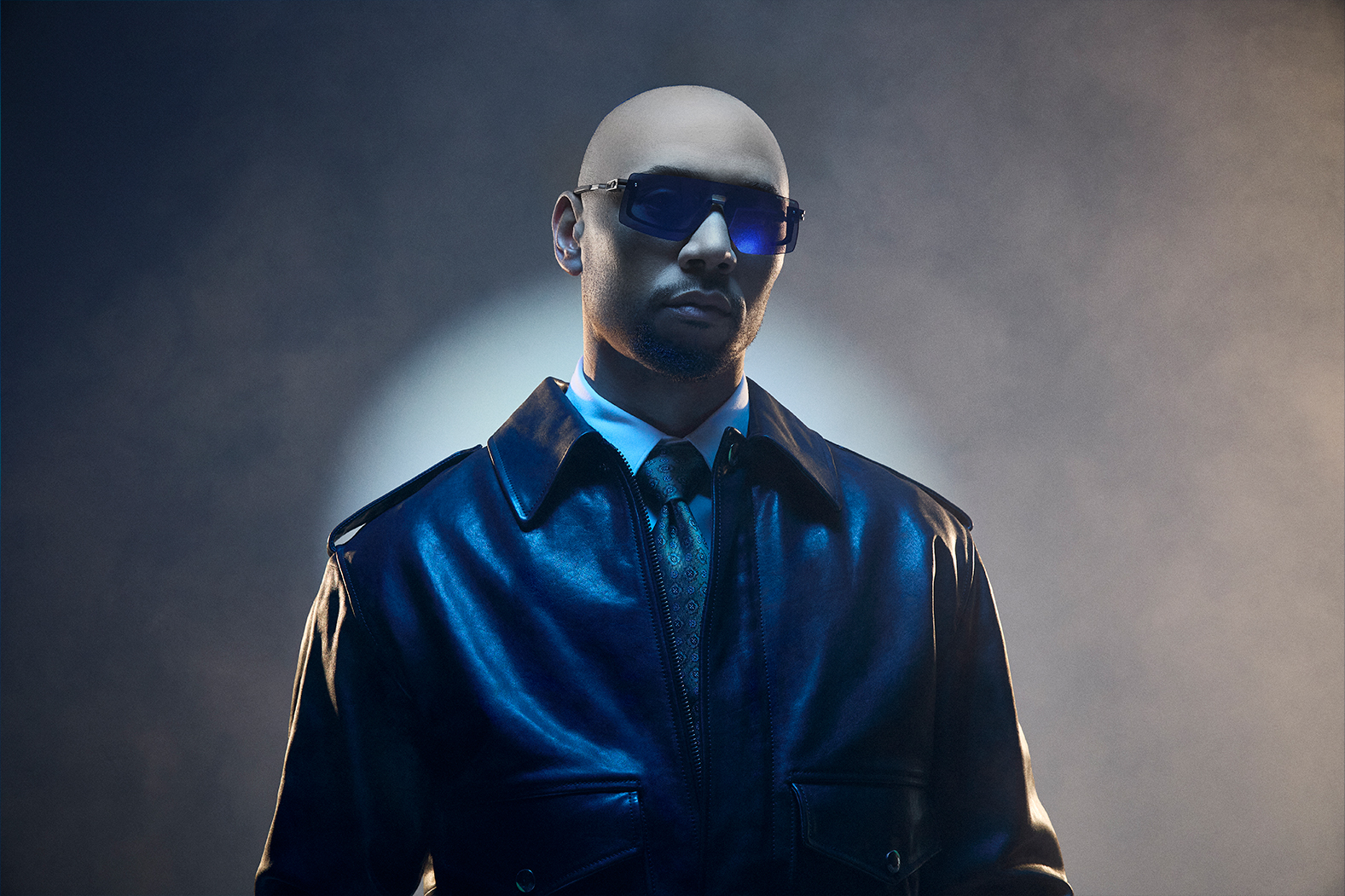 Man in sunglasses and leather jacket against a smoky background.