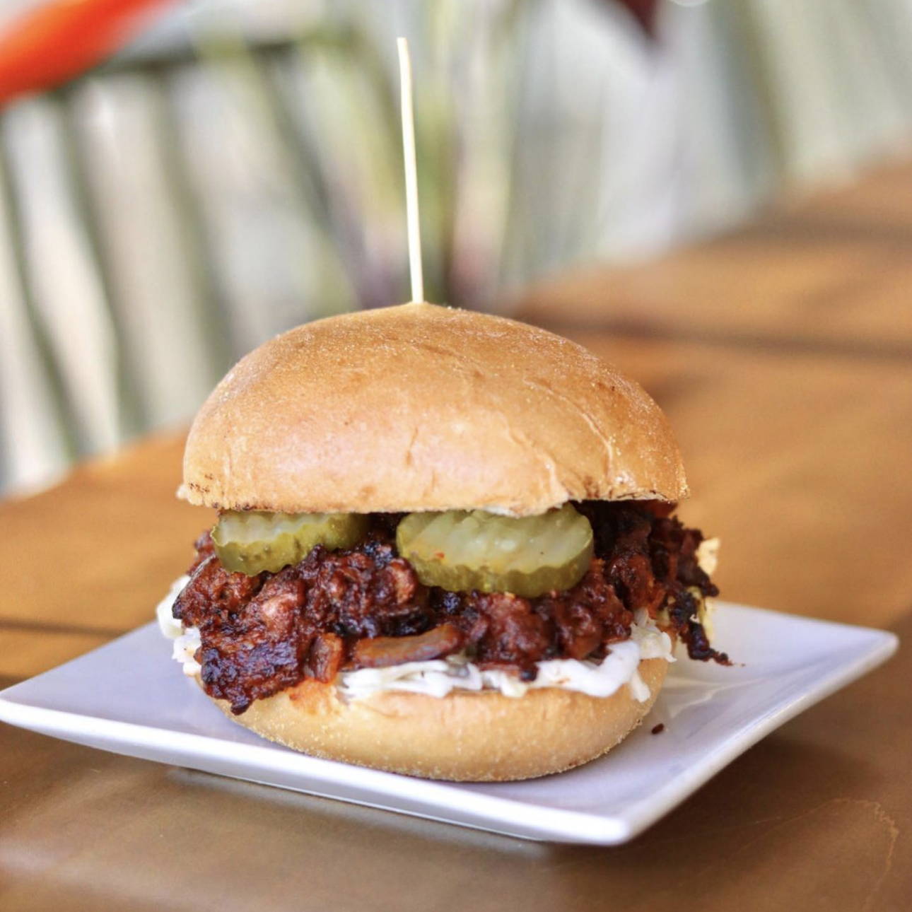 Harvest Shreds with BBQ sauce on a bun atop creamy cole slaw, topped with pickles. On a small square white plate.