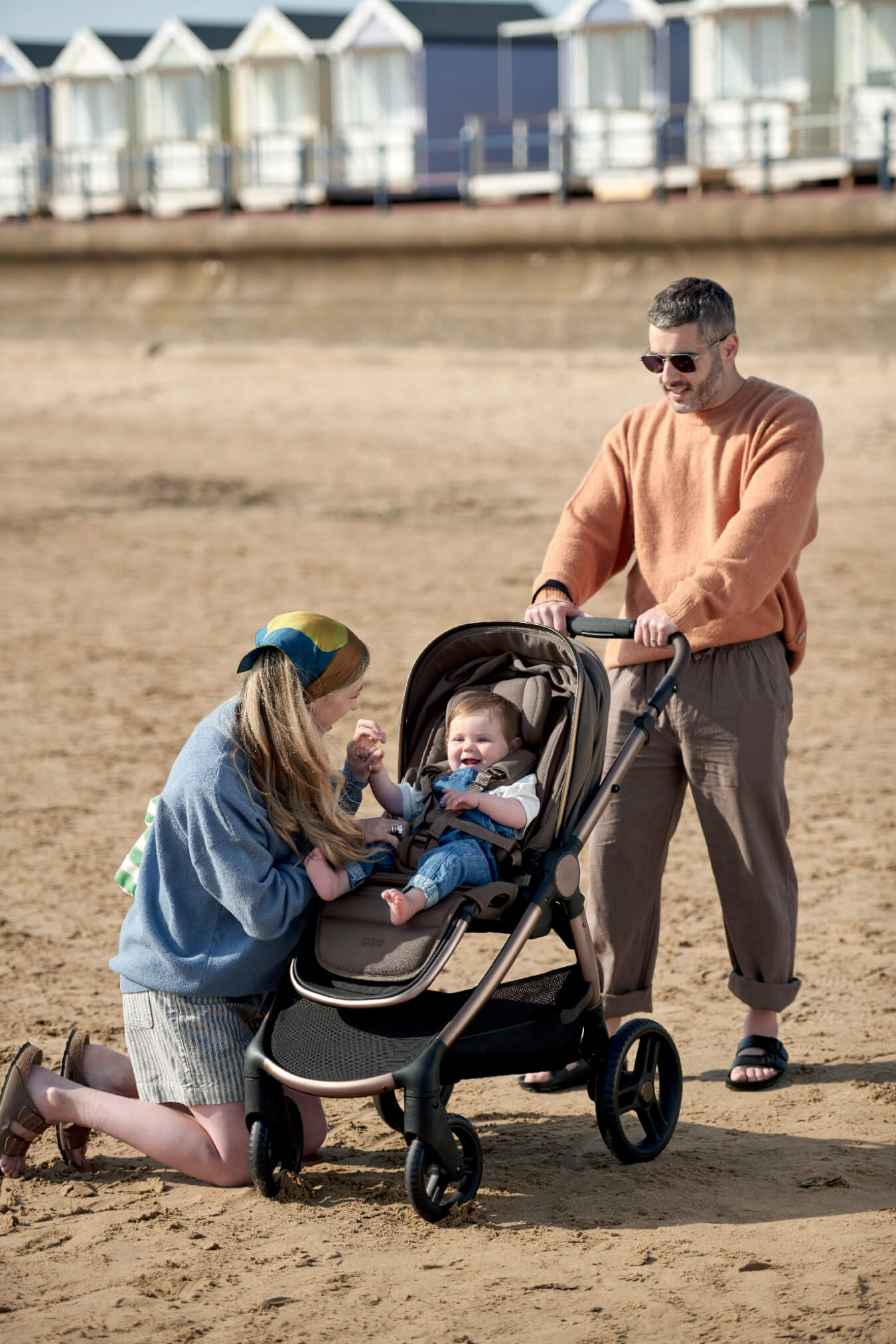 A woman interacts with a baby in a Ocarro 2 Cream Pushchair, while a man watches.