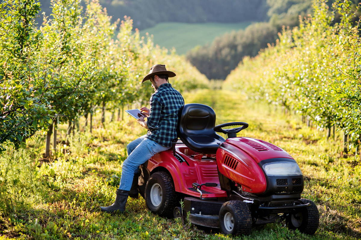 An mid-aged farmer resting on a small tractor in between rows of an orchard looking at a tablet
