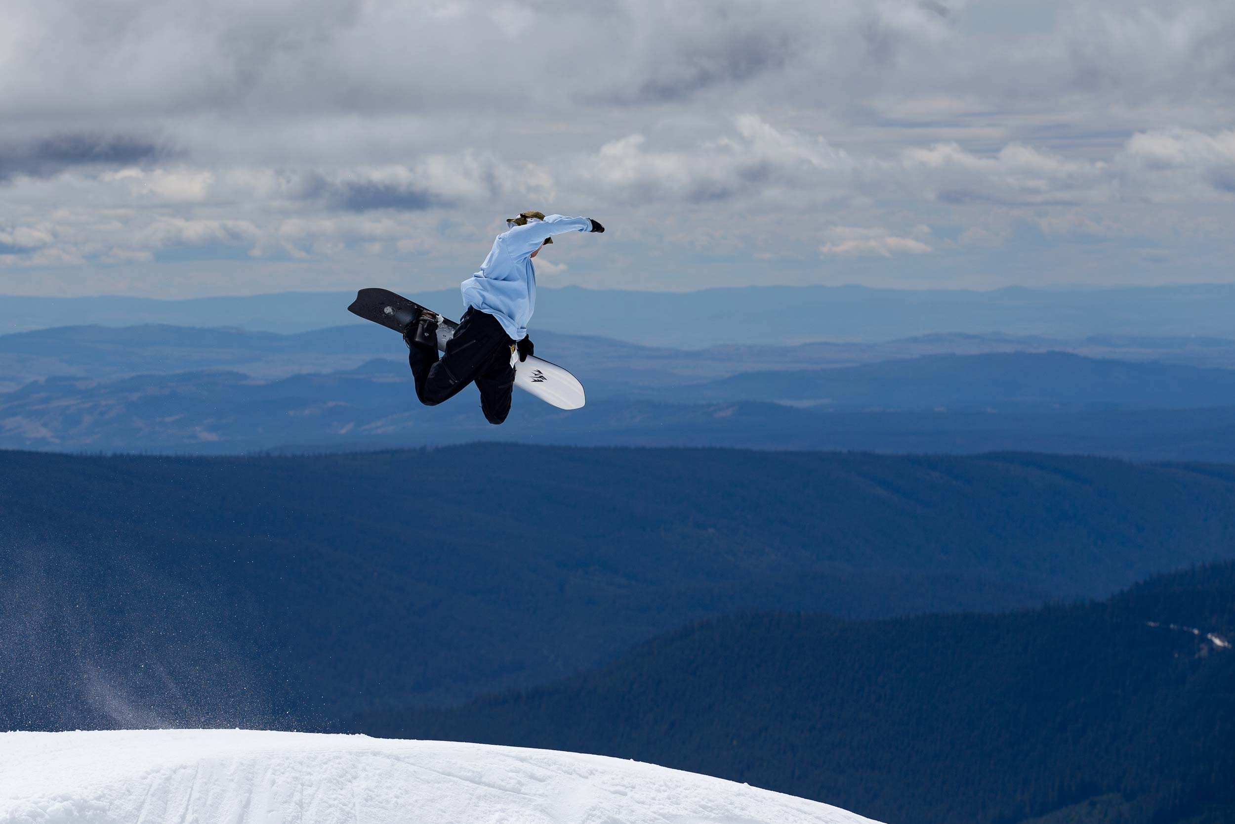 Snowboarder jumping mid-air against a mountainous landscape under a cloudy sky.