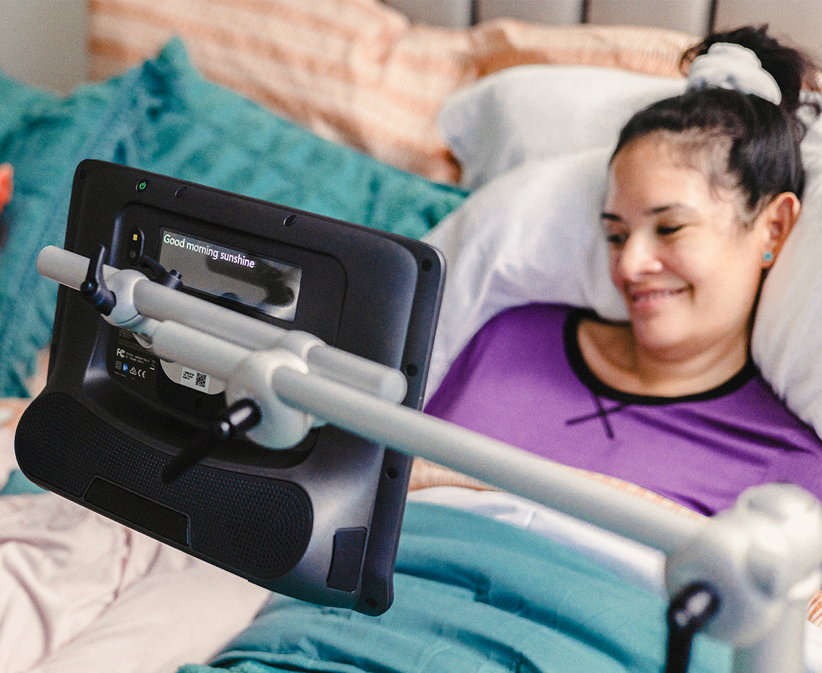 Floorstand mount for a Tobii Dynavox assistive communication device used by a woman in her bed