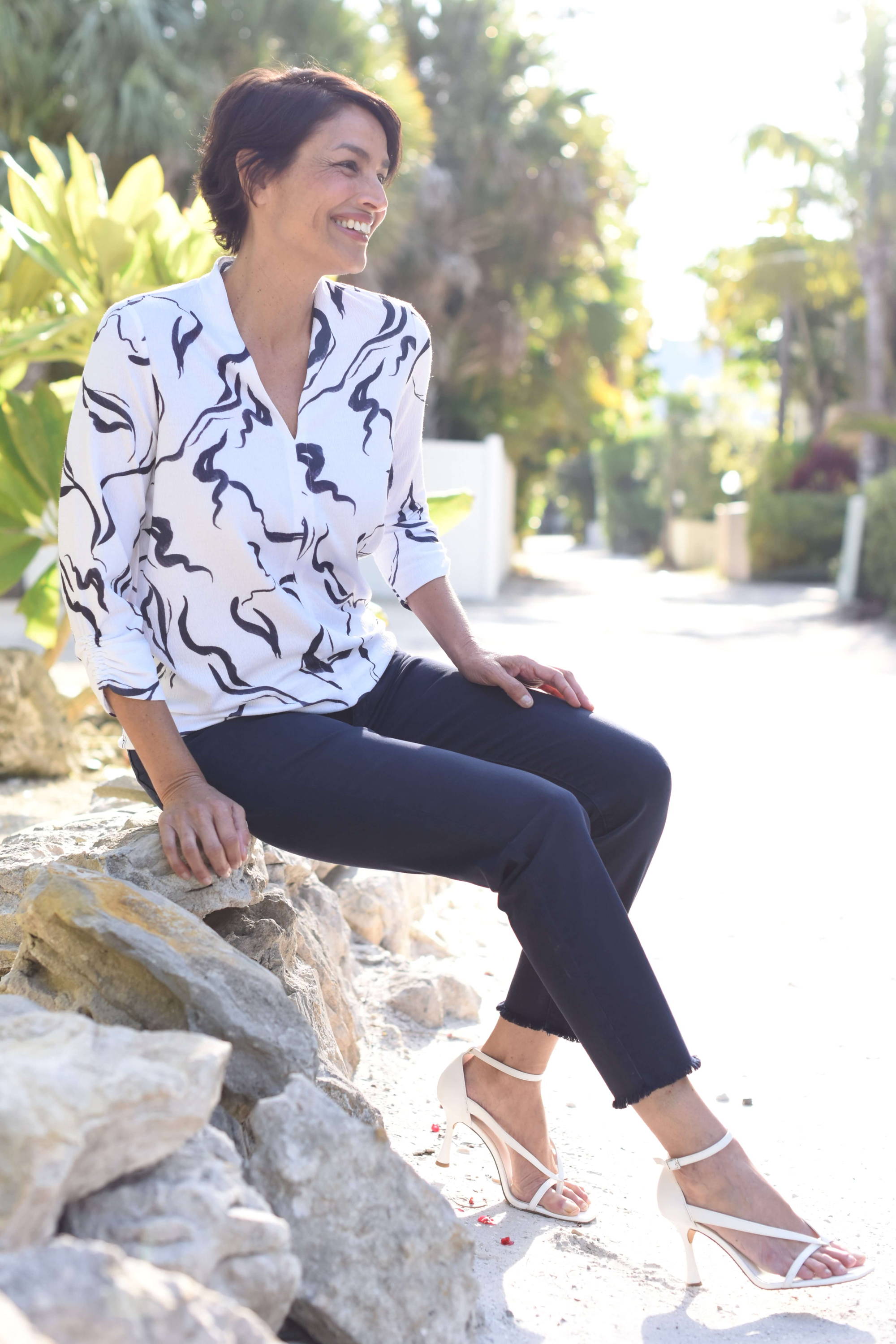 A smiling woman with short dark hair is wearing a printed top, ankle pants, and heels as she sits on a rock near the ocean