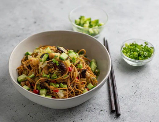 A bowl of noodles topped with vegetables and green onions, chopsticks beside it.