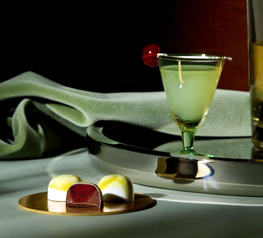 A cocktail glass and colorful desserts on a reflective surface.