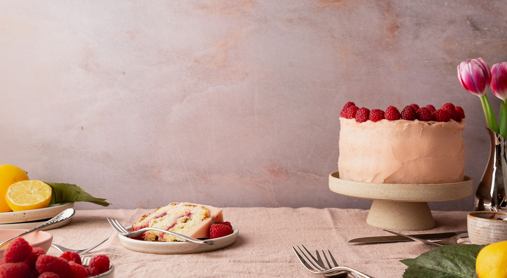 A cake with raspberries, a slice, lemons, and flowers on a table.