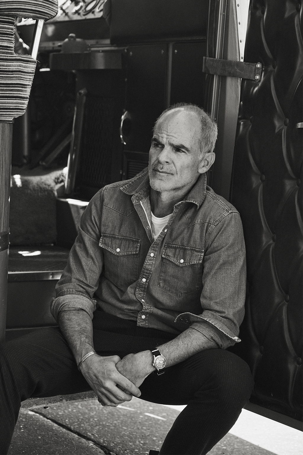 Michael Kelly Sitting on the steps of a vintage bus wearing the Ennis Selvedge Denim Western Shirt in Medium Wash and the Rivay Italian Wool Suit in Charcoal Herringbone Trousers