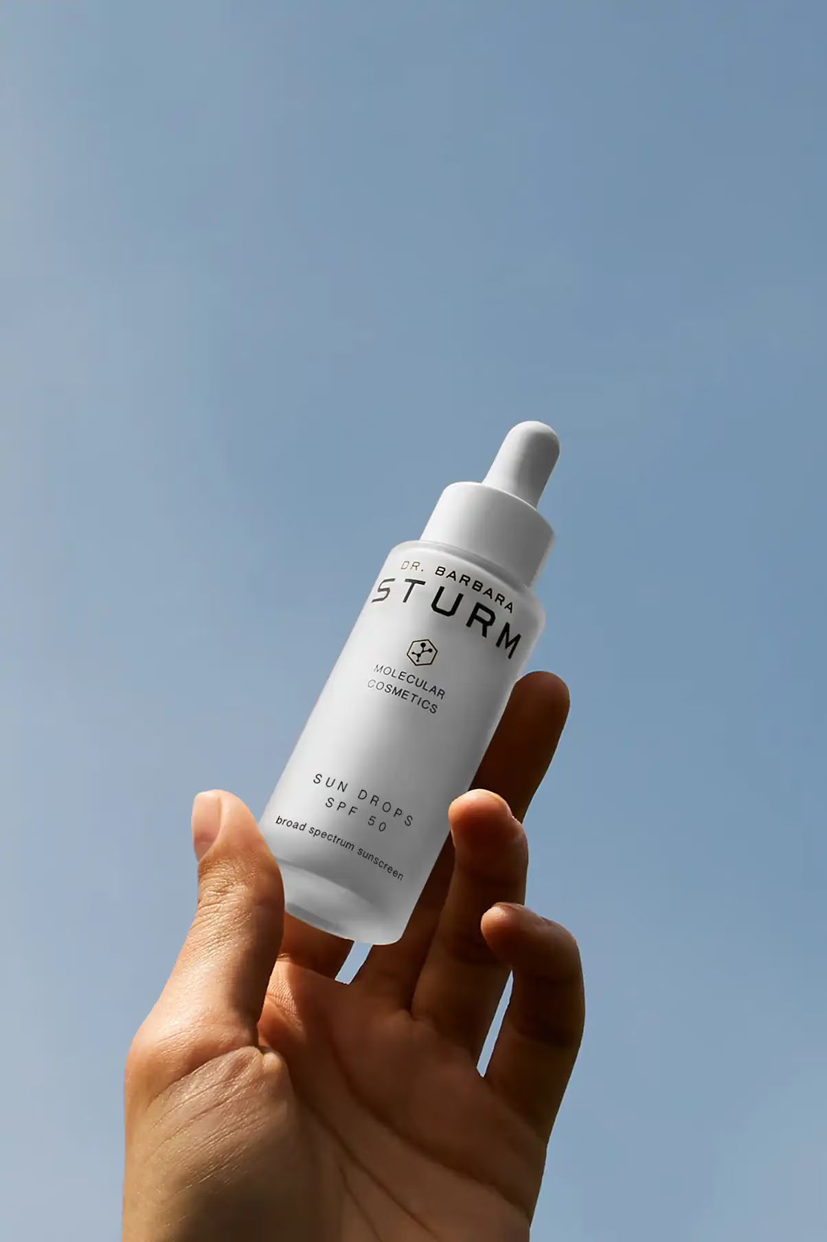 A hand holding a skincare serum bottle against a clear blue sky.