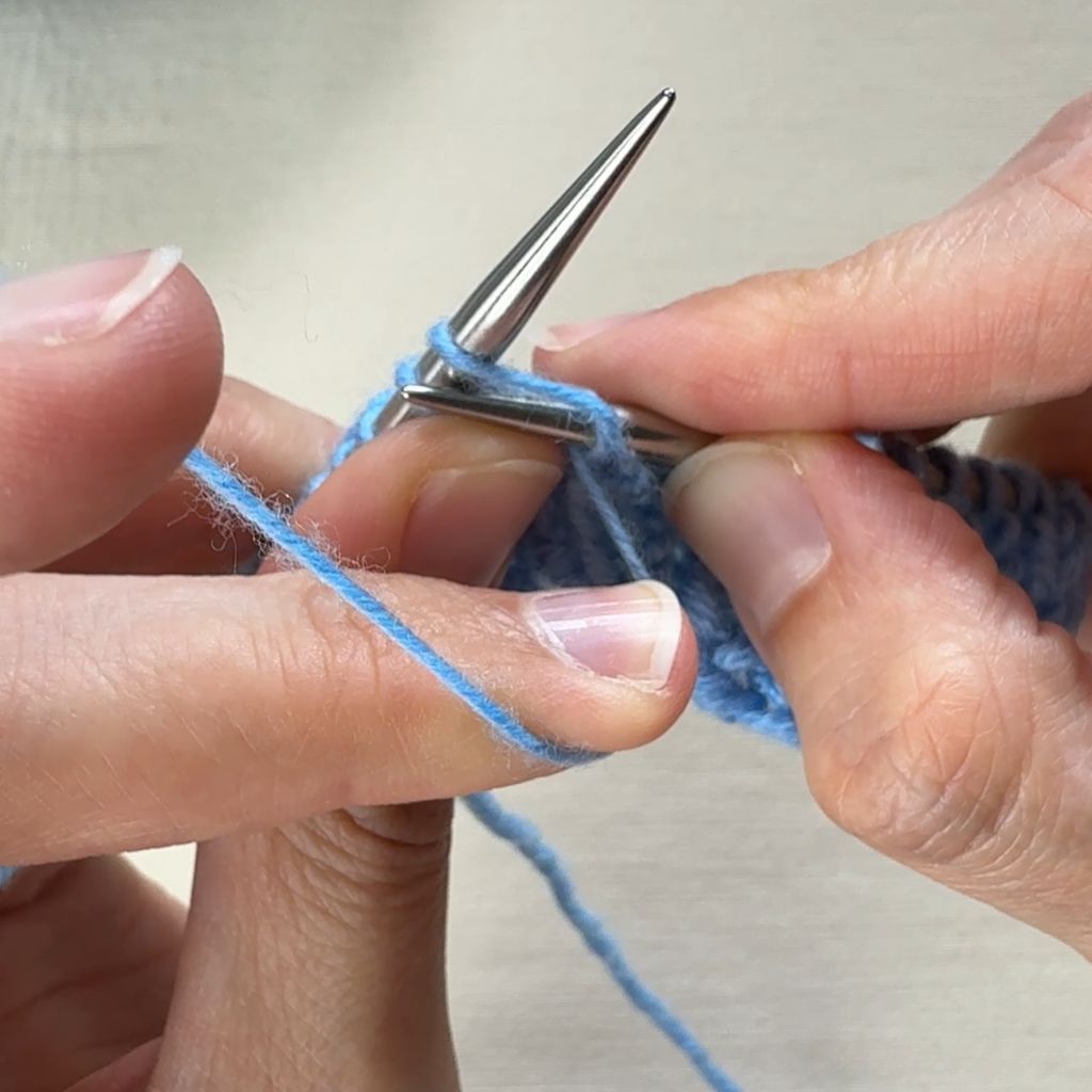 Detailfoto, wie eine linke Masche gestrickt wird. Um die rechte Nadel wird eine Schlaufe gebildet und der Faden anschließend durch die Masche gezogen.