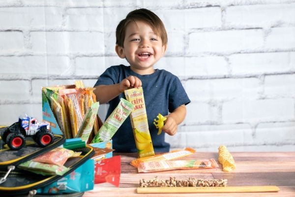 Smiling child holding snacks with toys and a backpack on a table.