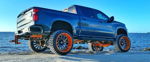 Rear three-quarter Mauricio's lifted blue Chevrolet Silverado RST on sandy shore with orange accents photo 1