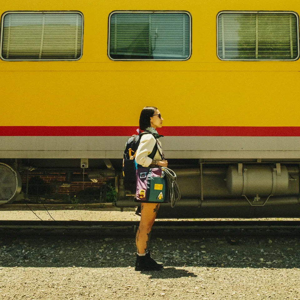 Person standing beside a yellow train, wearing a backpack and colorful outfit.