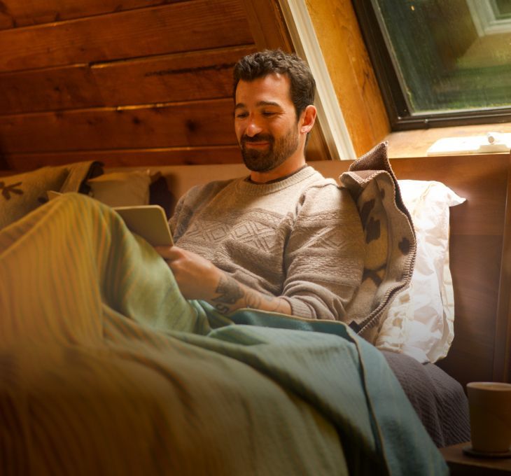 Man sitting on a bed, reading a tablet, smiling. Cozy atmosphere.