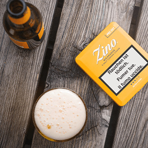 Die gelbe Dose der Zino Nicaragua Half Corona-Zigarren auf einem Holztisch neben einem gefüllten Bierglas.