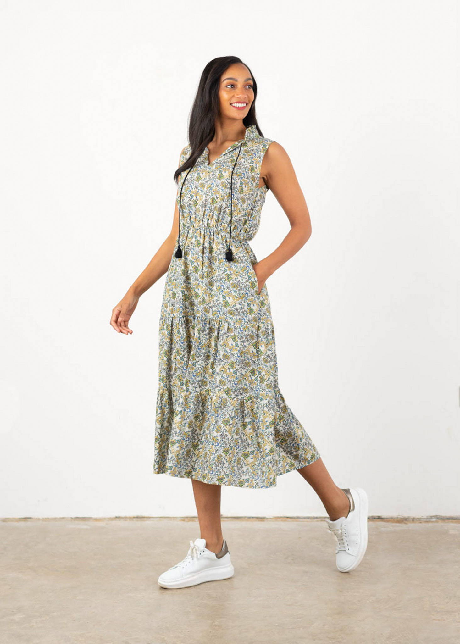 A model wearing a sleeveless midi dress with a floral patter and tassles with white trainers