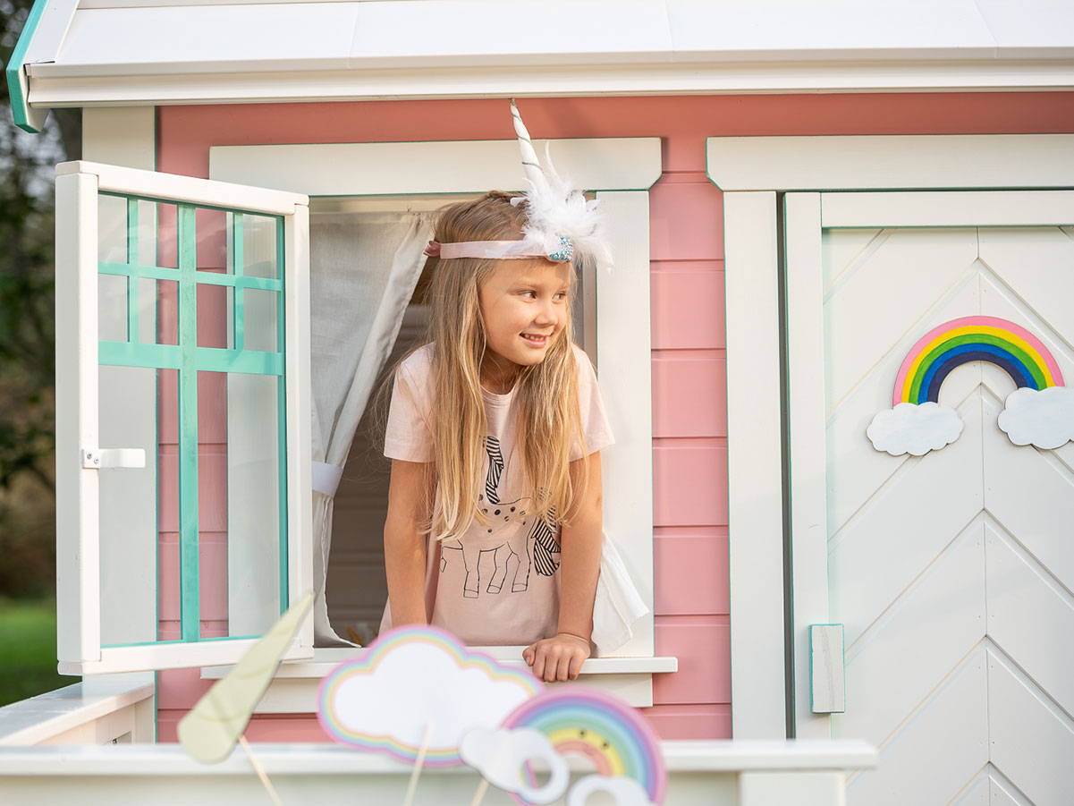  One girl looks out of the window of a decorated kids outdoor playhouseby WholeWoodPlayhouses