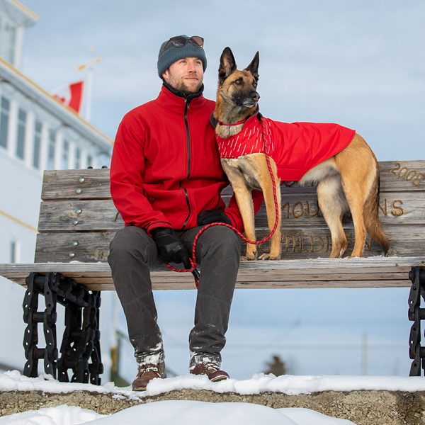 Person in red coat sits on bench with dog in matching red coat.