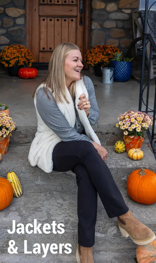 Woman sitting on steps surrounded by pumpkins and flowers, wearing a cozy outfit.