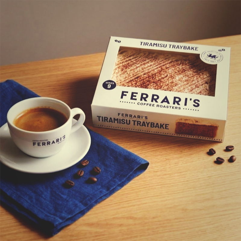 Ferraris Coffee Tiramisu Traybake in branded packaging beside a cup of Ferraris espresso, with coffee beans and a blue napkin on a wooden table