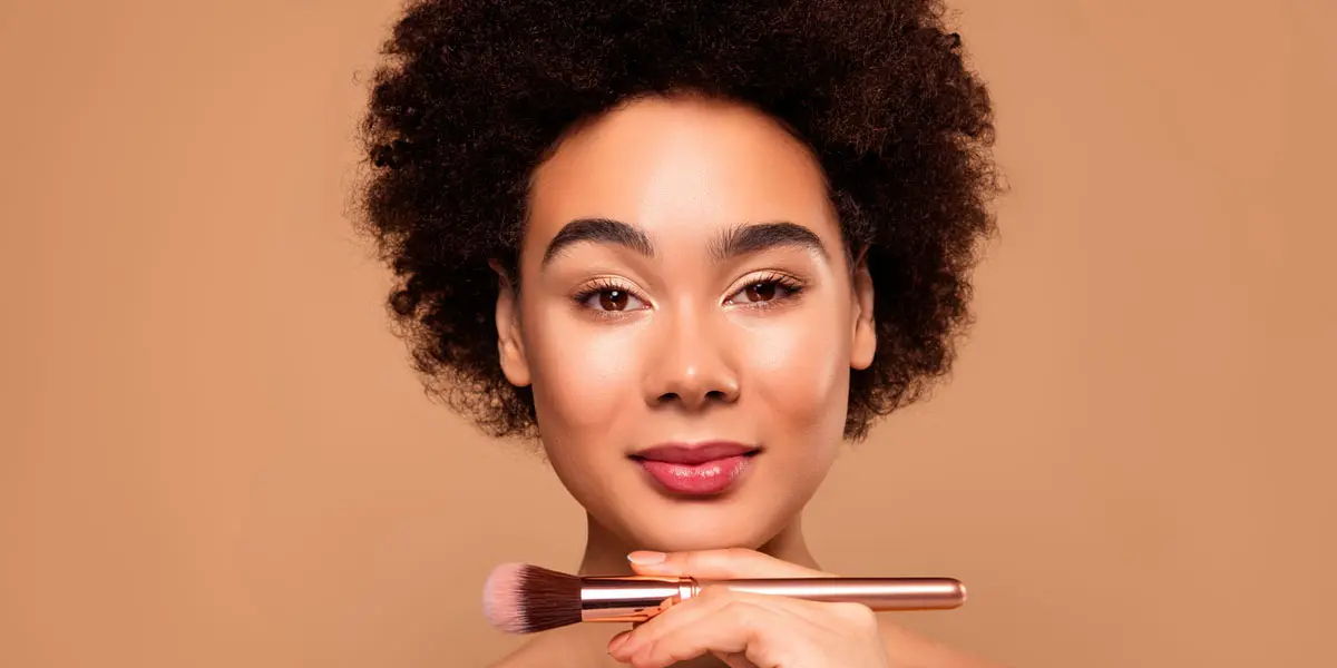 Woman with curly hair holding a makeup brush, smiling against a beige background.