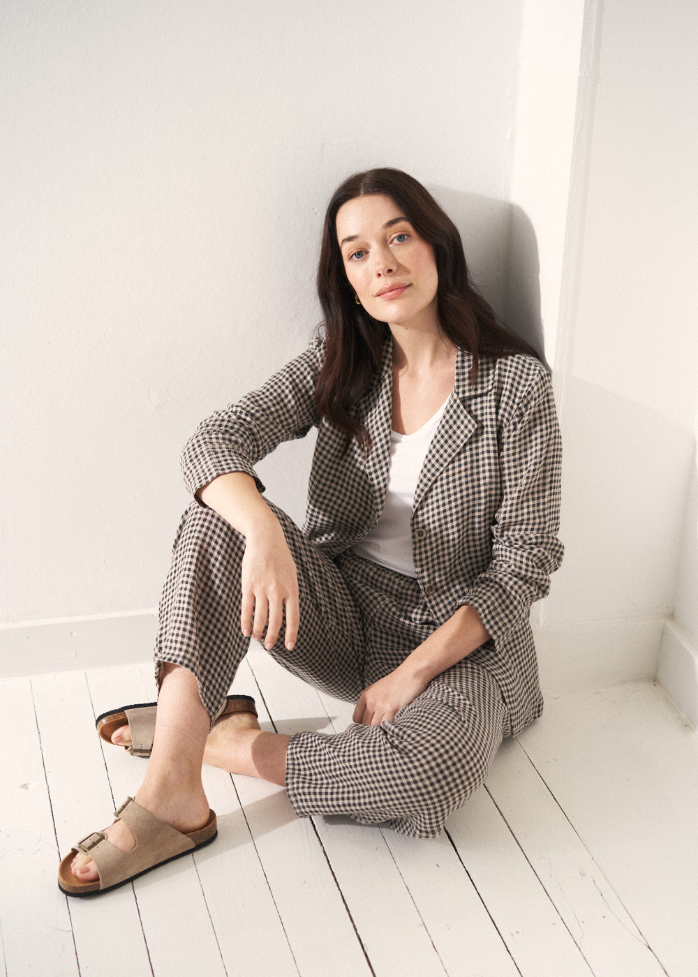A woman models a grey and black checked linen jacket over a white top, paired with matching barrel-leg checked linen trousers. She sits in front of a beige background.