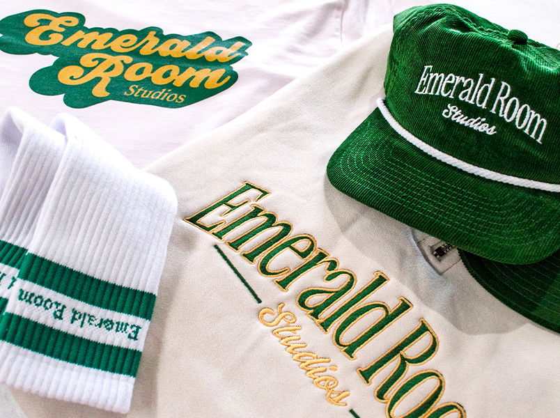Green and white Emerald Room Studios embroidered cord caps, woven socks, and screenprinted t-shirts and embroidered sweatshirt with the Emerald Room logo in green and gold