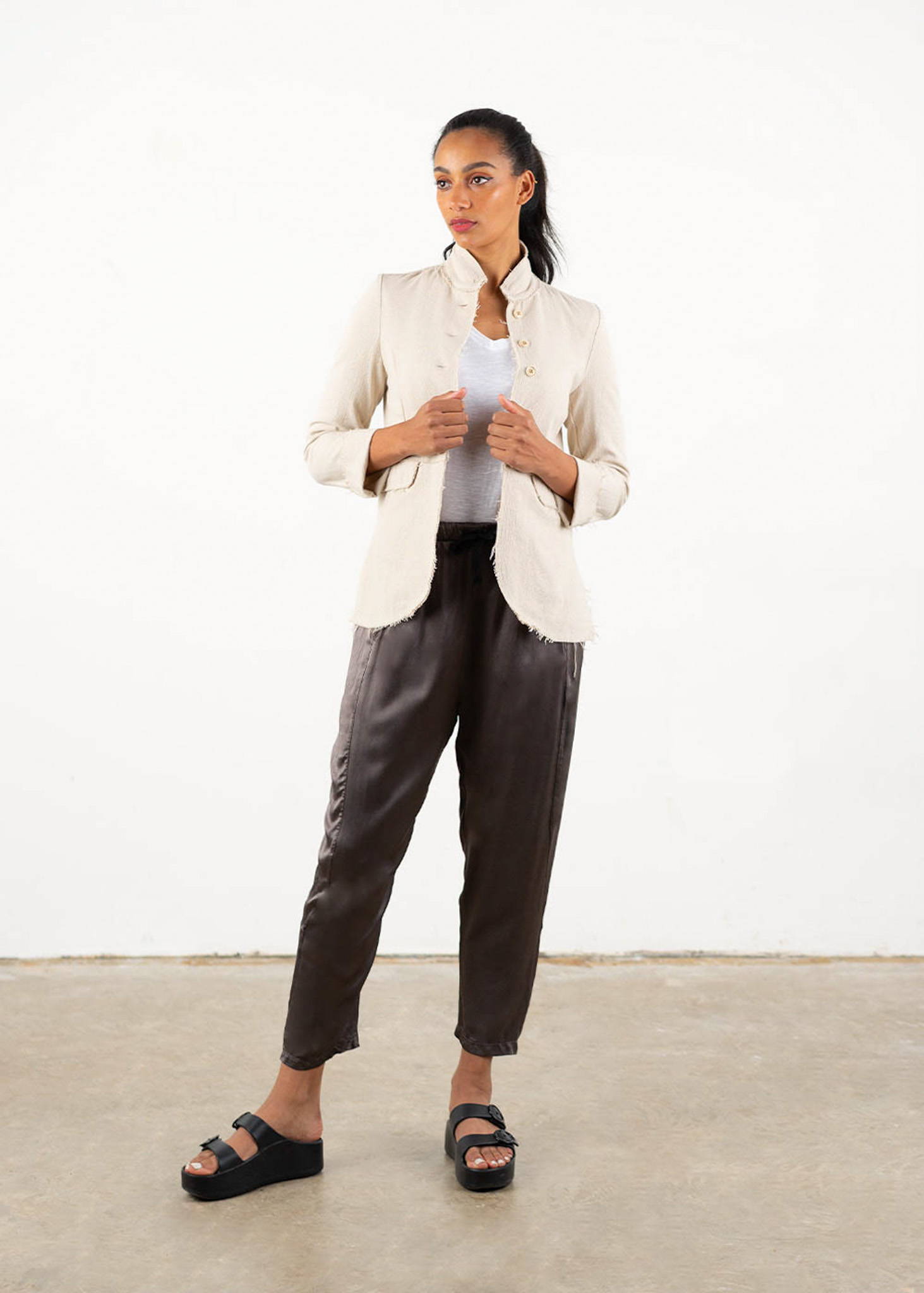A model wearing a off white, raw hemmed fitted jacket over a white top, dark grey satin trousers and black platform slides.