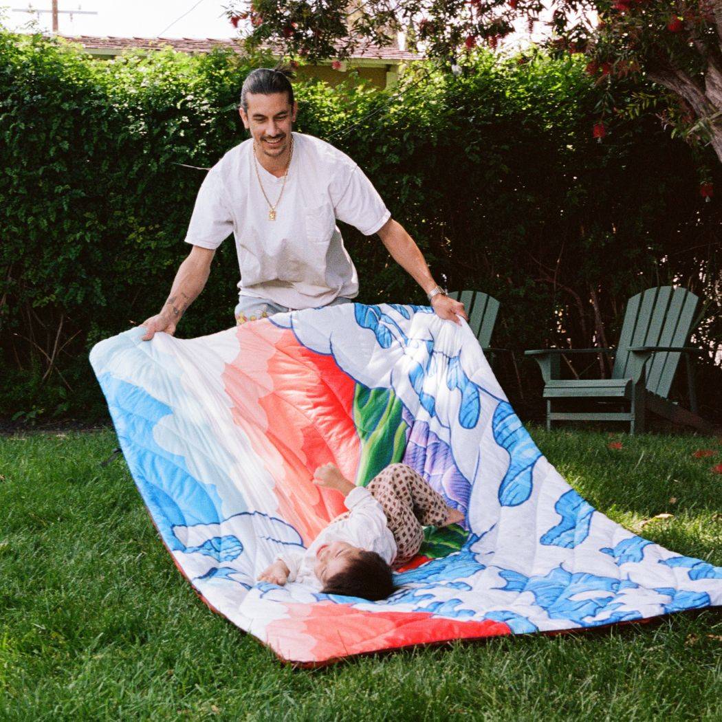 Aaron Kai sharing a playful moment with his son, who rides the a Rumpl Original Puffy Blanket across the grass, laughing. 