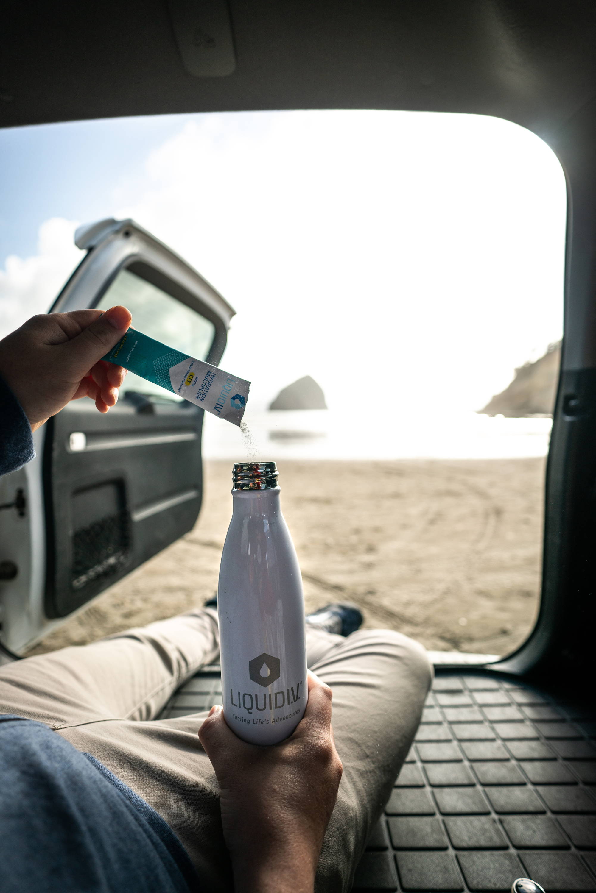 Active outdoorsy adult pours Liquid IV's electrolyte powder into his water bottle from the back of his van.