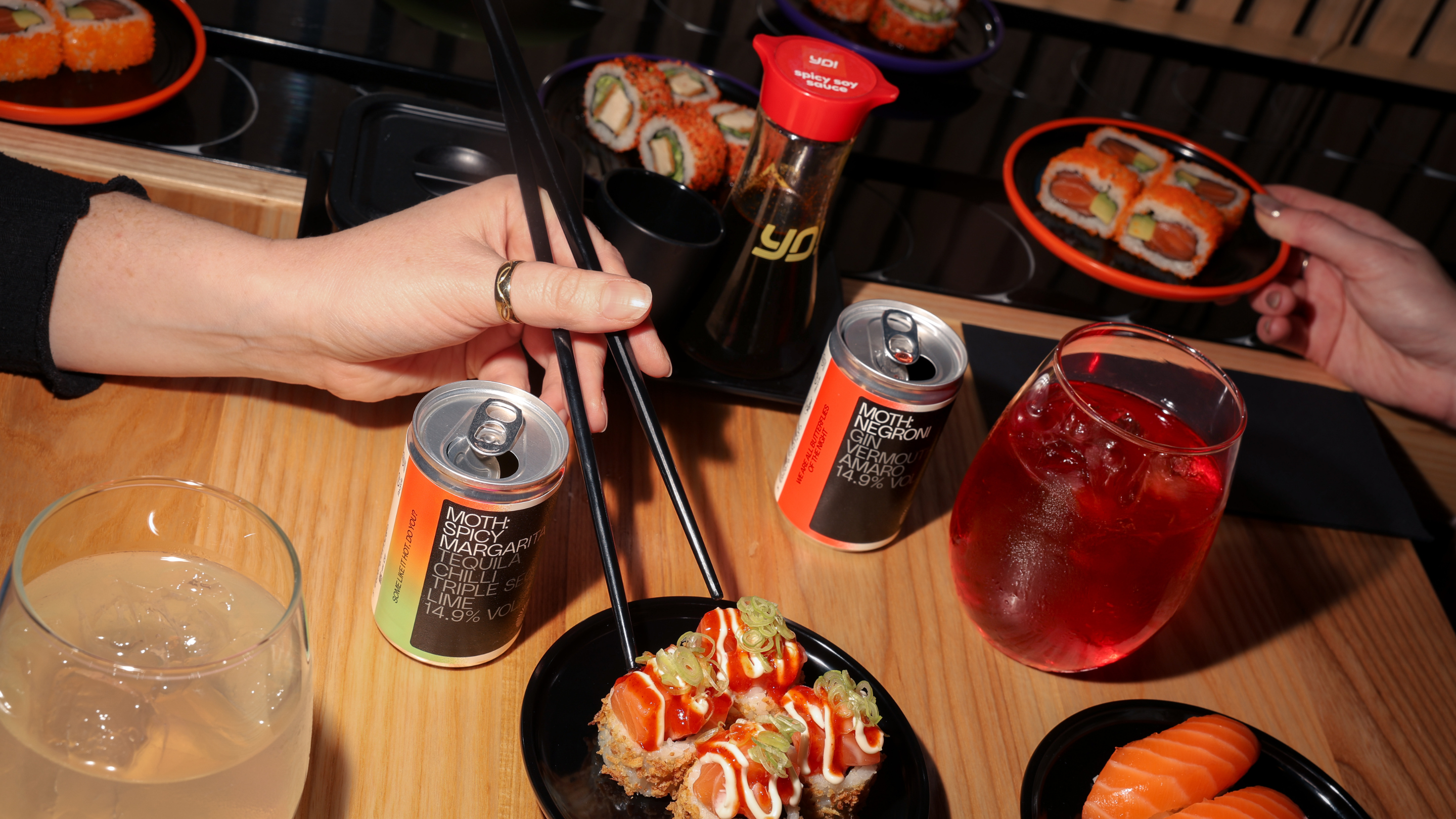 Hands holding sushi, drinks, and chopsticks on a wooden table.