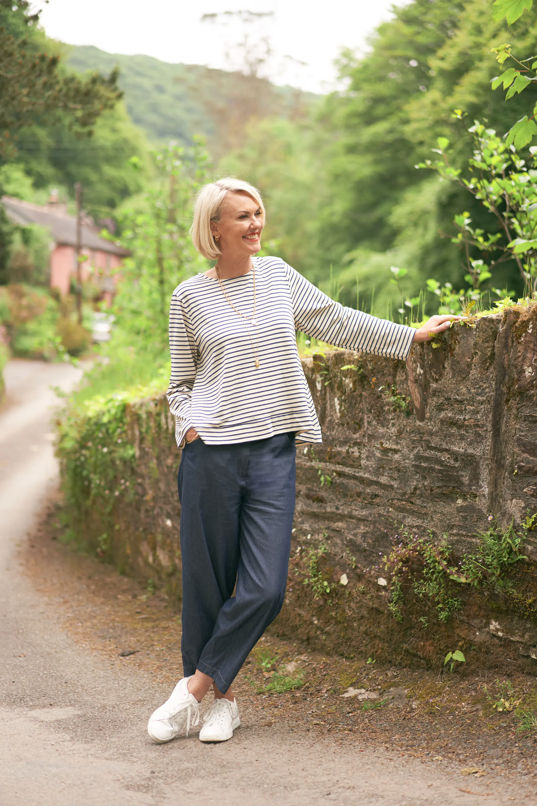 Woman wearing a long sleeve striped top with dark blue straight leg jeans and white pumps, leant against a stone wall