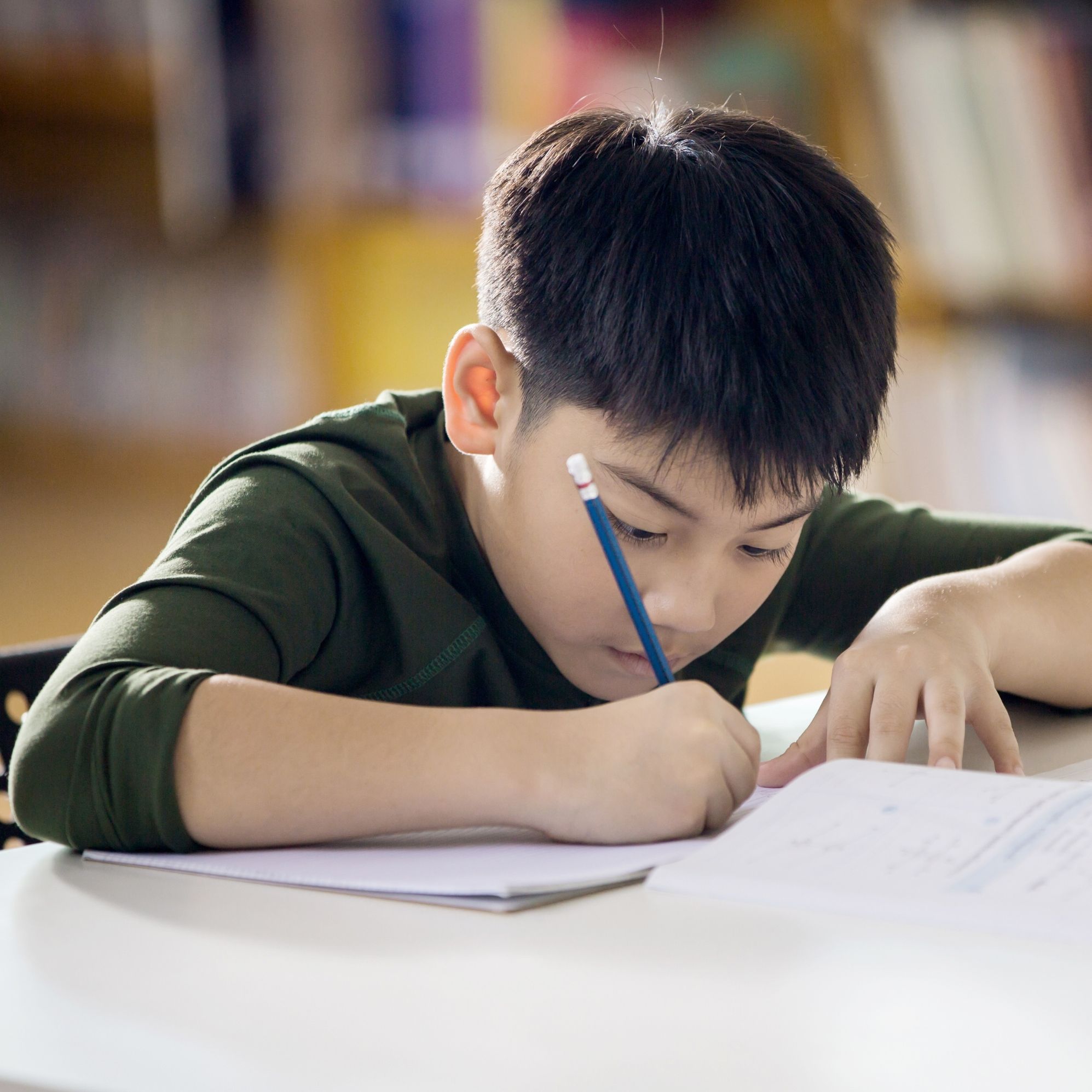 A boy writing in a notebook at a table in a library.