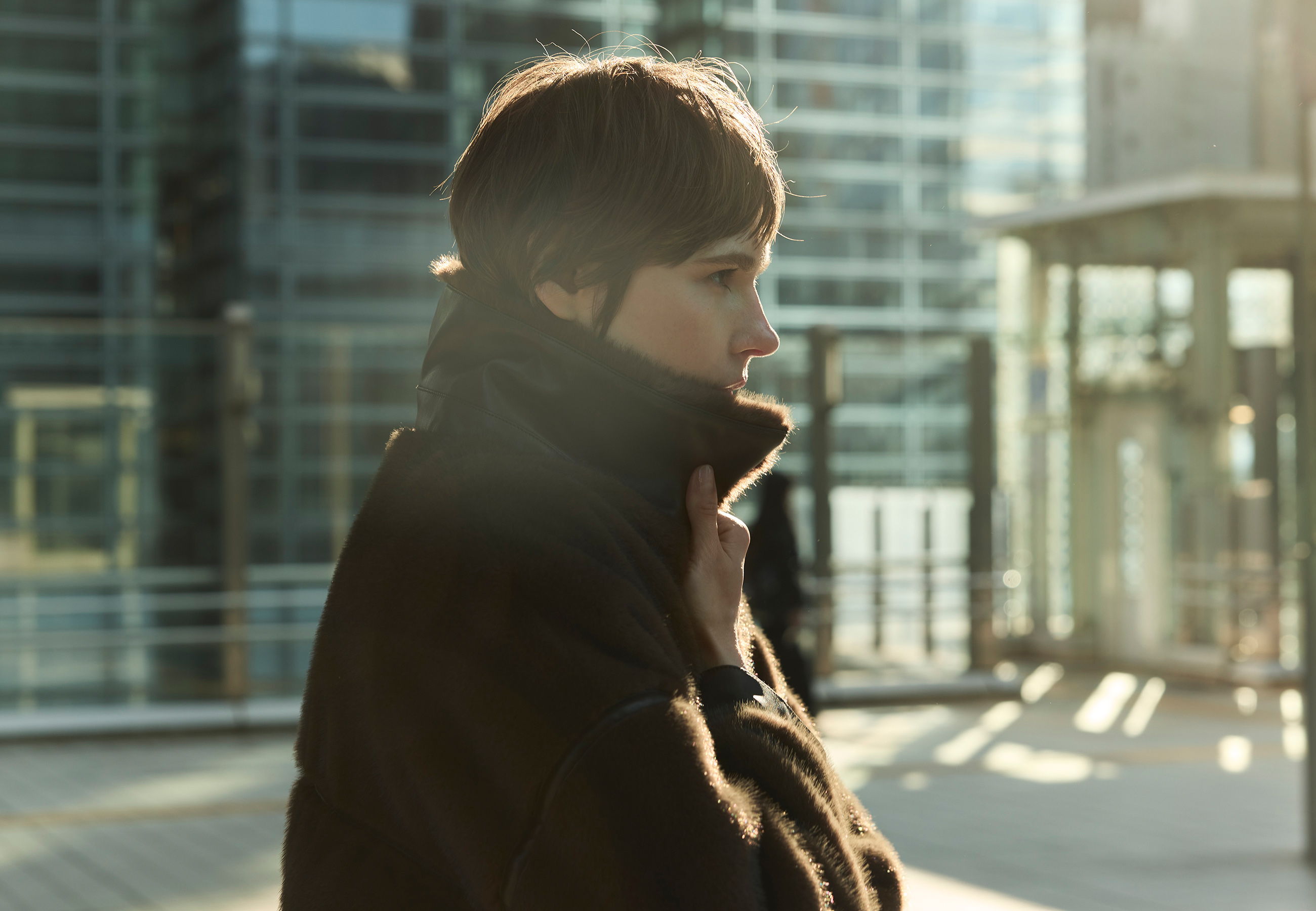 Person in a coat standing outdoors with sunlight and modern buildings in background.