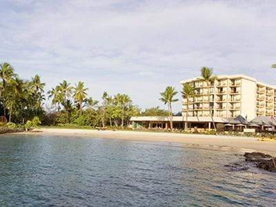 Exterior photo of hotel. You can see the ocean and palm trees on the shore. 
