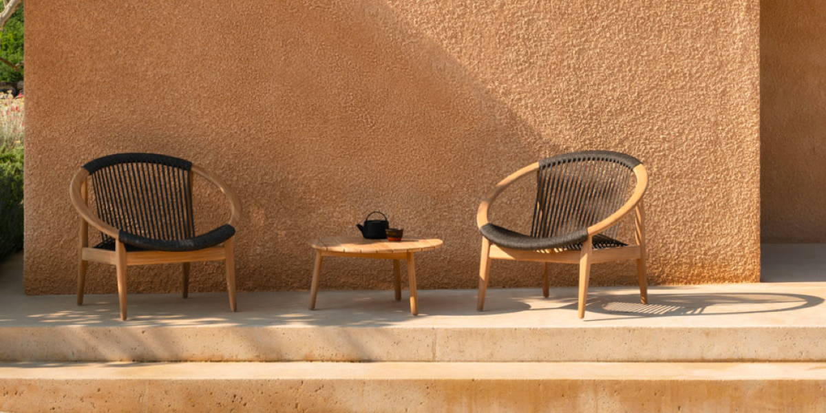 Two Frida outdoor lounge chairs made with a circular teak frame and dark woven seat are arranged in front of a stucco wall.