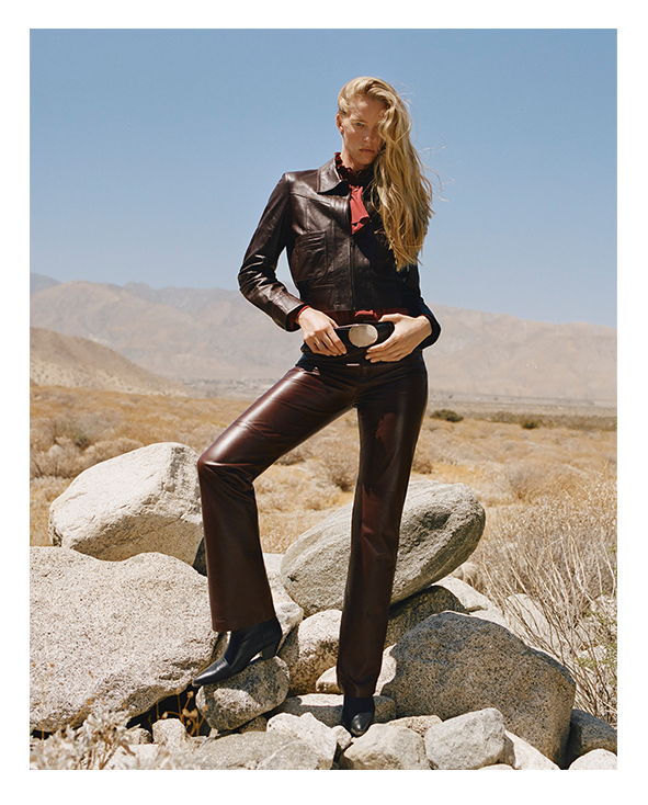 A person in a leather outfit stands on rocks in a desert landscape.