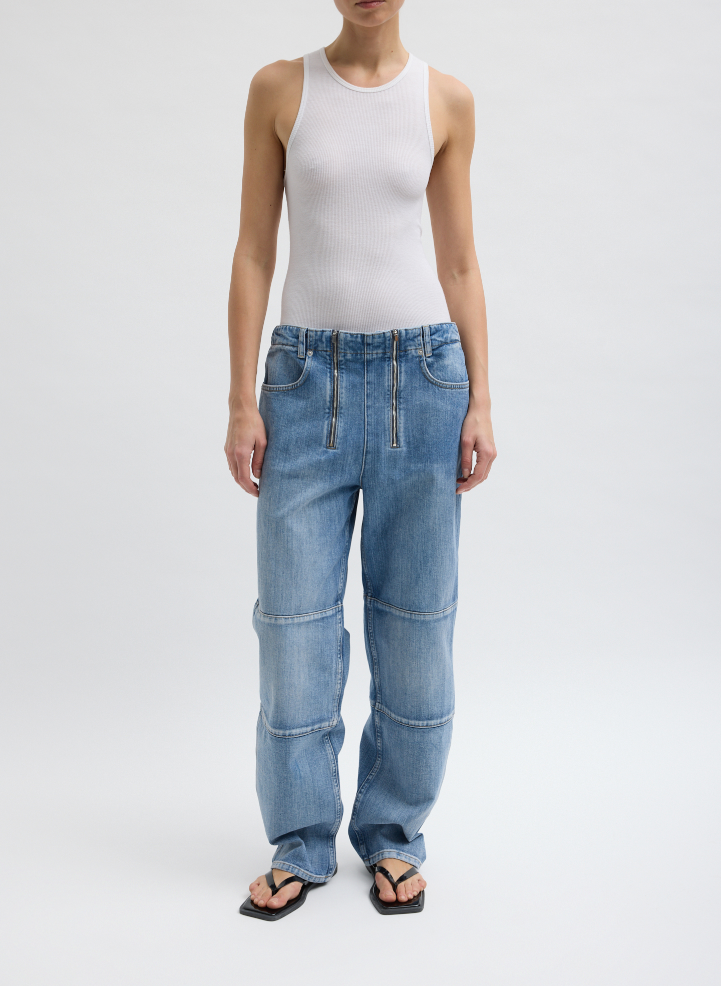 Person wearing a white tank top and baggy blue jeans, standing on a plain background.