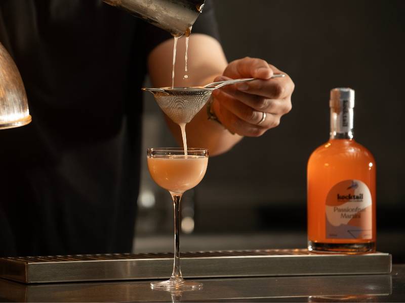 Bartender serving a Passionfruit martini into a coupe glass through a sieve on a bar