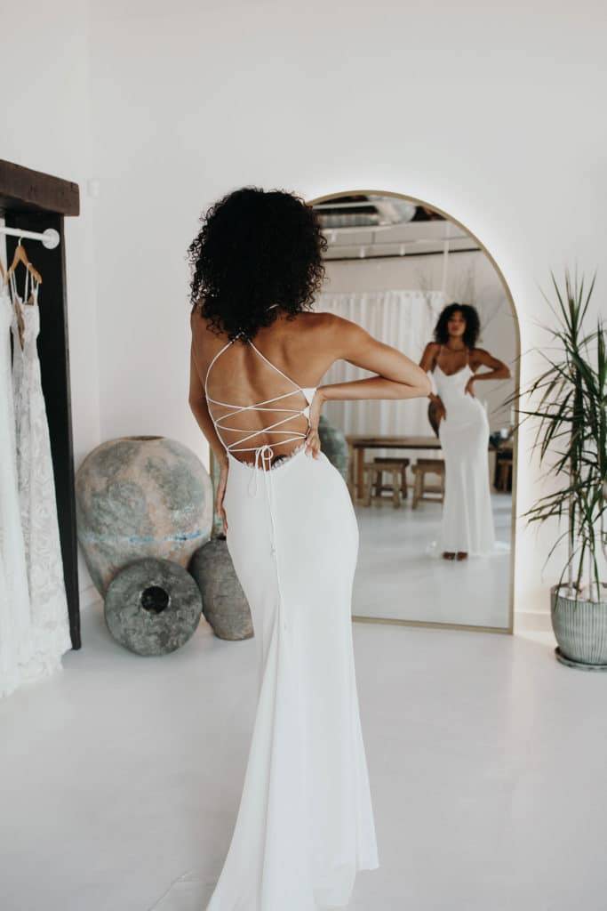 Mariée portant une robe en dentelle de soie à nouer dans le dos devant un miroir en arc de cercle dans une pièce blanchie à la chaux.