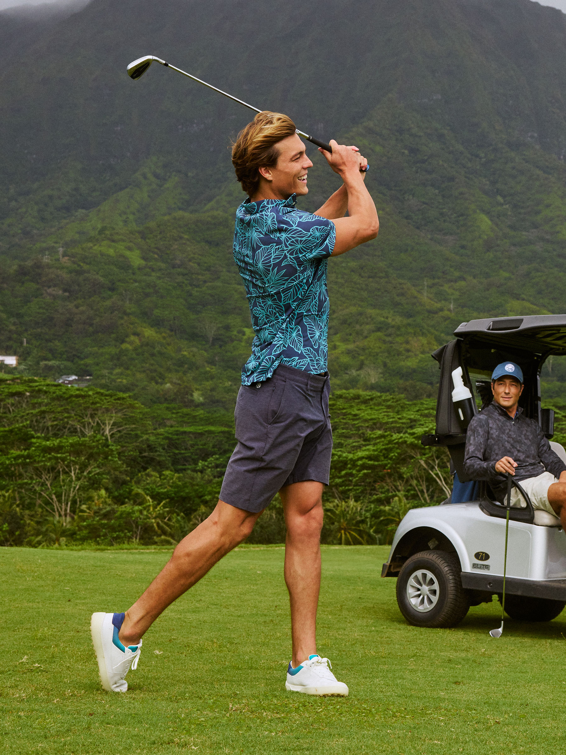 Man swinging a golf club on a course with mountains in the background.