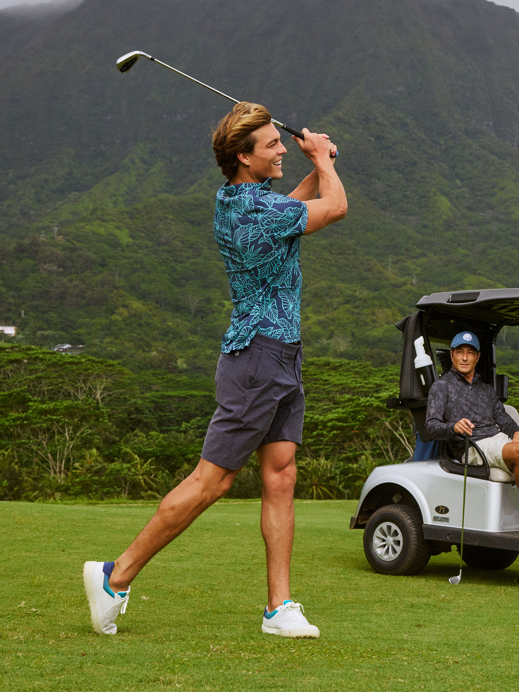Man swinging a golf club on a course with mountains in the background.
