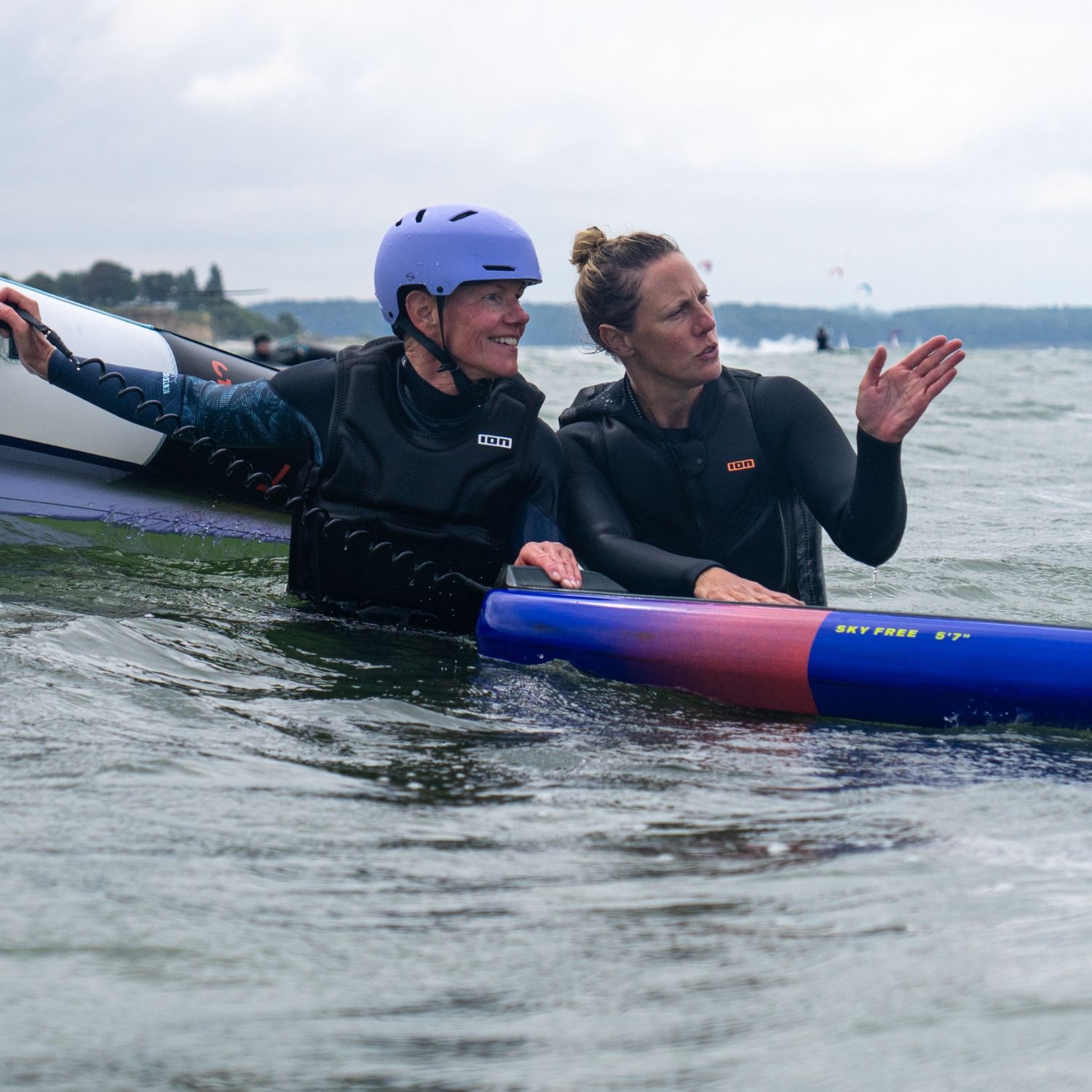 Two people in wetsuits, sitting on a surfboard in the water.