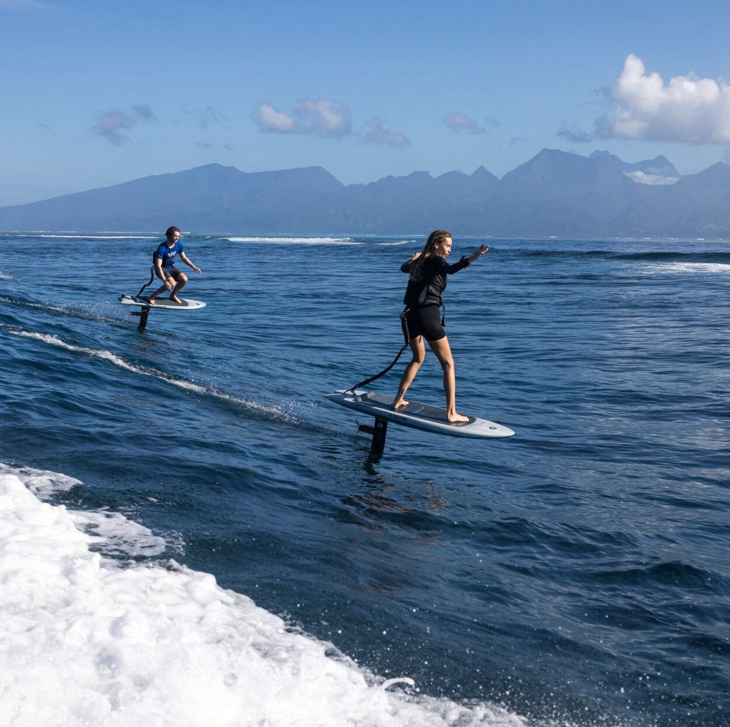 Two people surfing on hydrofoils in the ocean with mountains in the background.