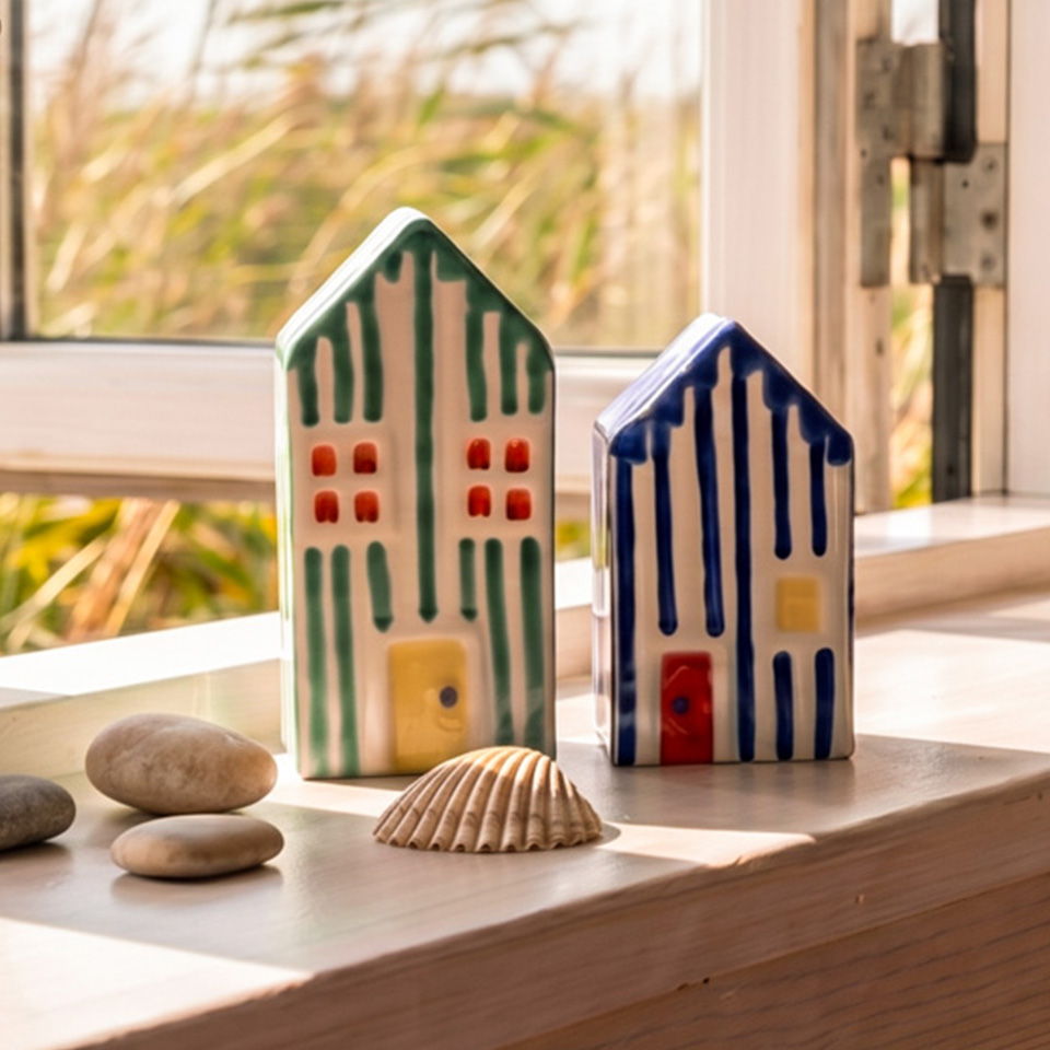 Two ceramic house figurines beside pebbles and a seashell on a windowsill.