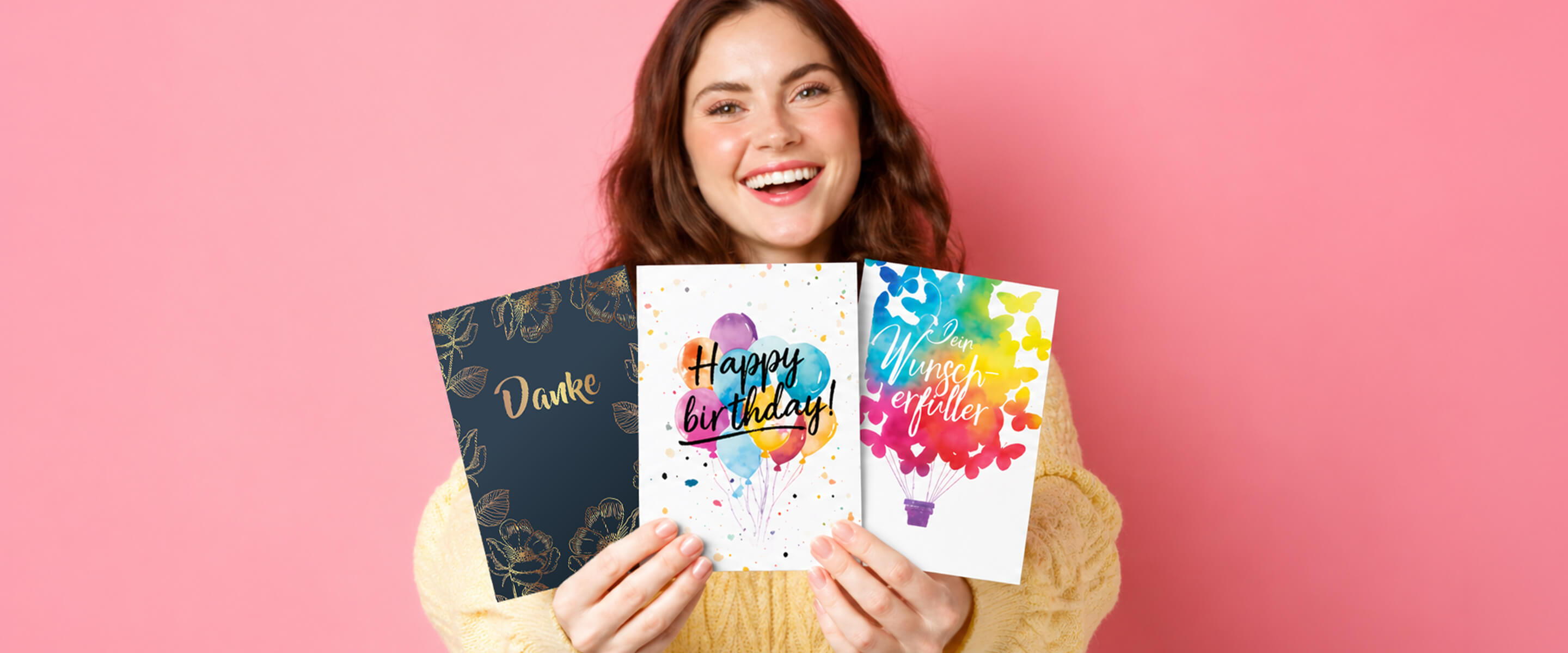 Smiling woman holding colorful greeting cards against a pink background.