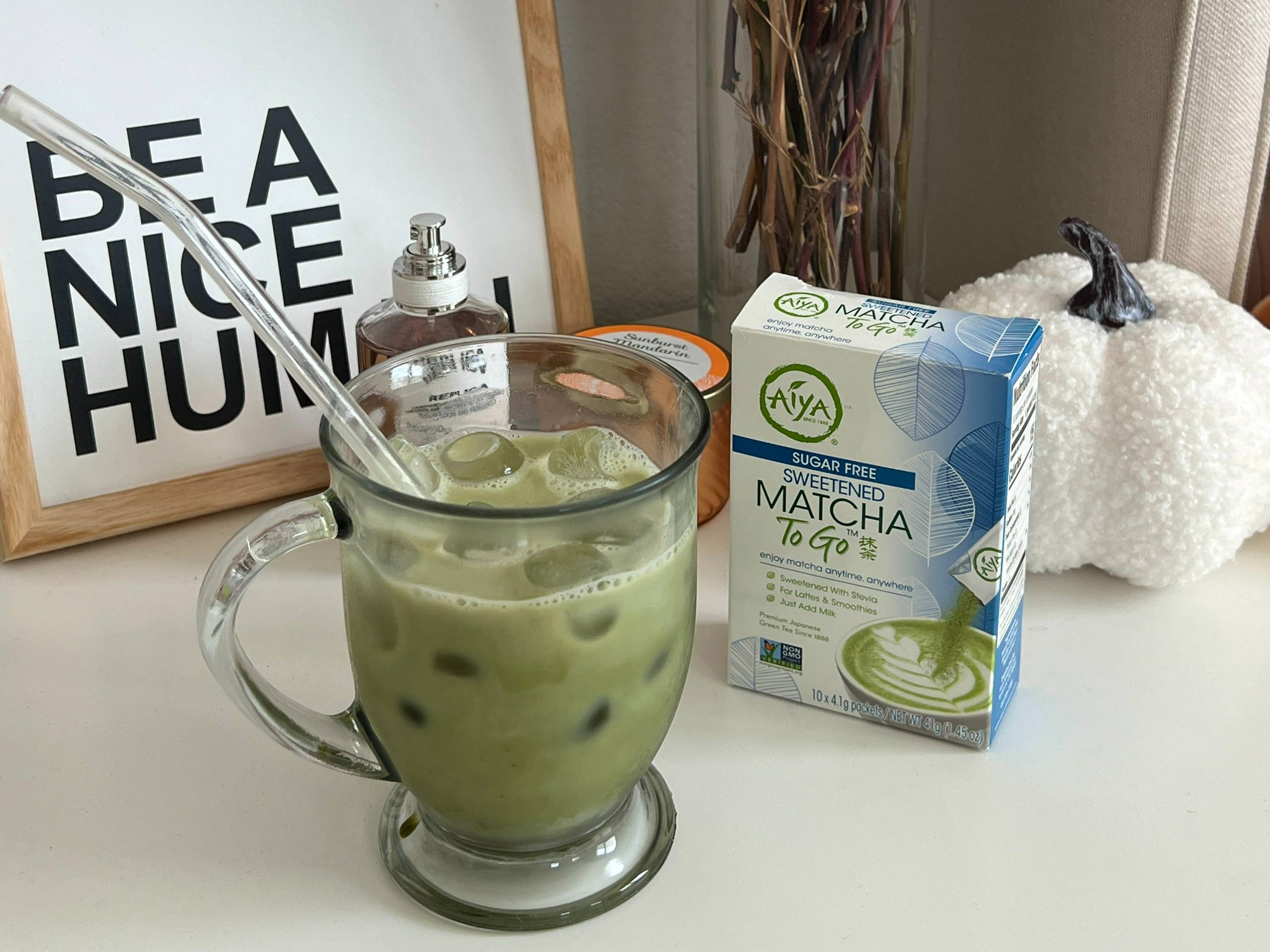 A glass of iced matcha next to decorative items and a carton of Sugar Free Sweetened Matcha To Go.