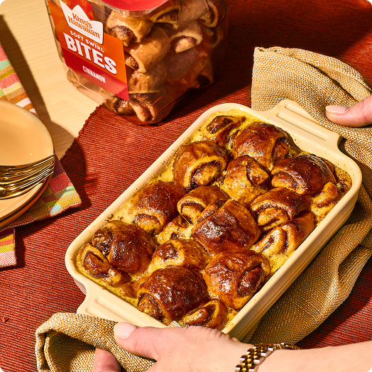 Baked cinnamon rolls in a dish, hands holding a towel, snack packaging in background.