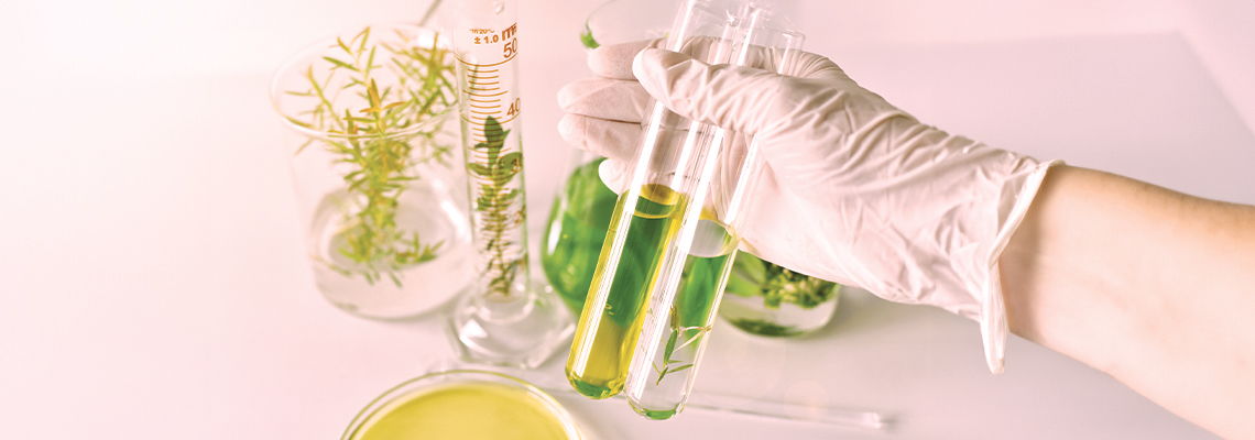 Hand in glove holding test tubes with green liquid and plants in background.