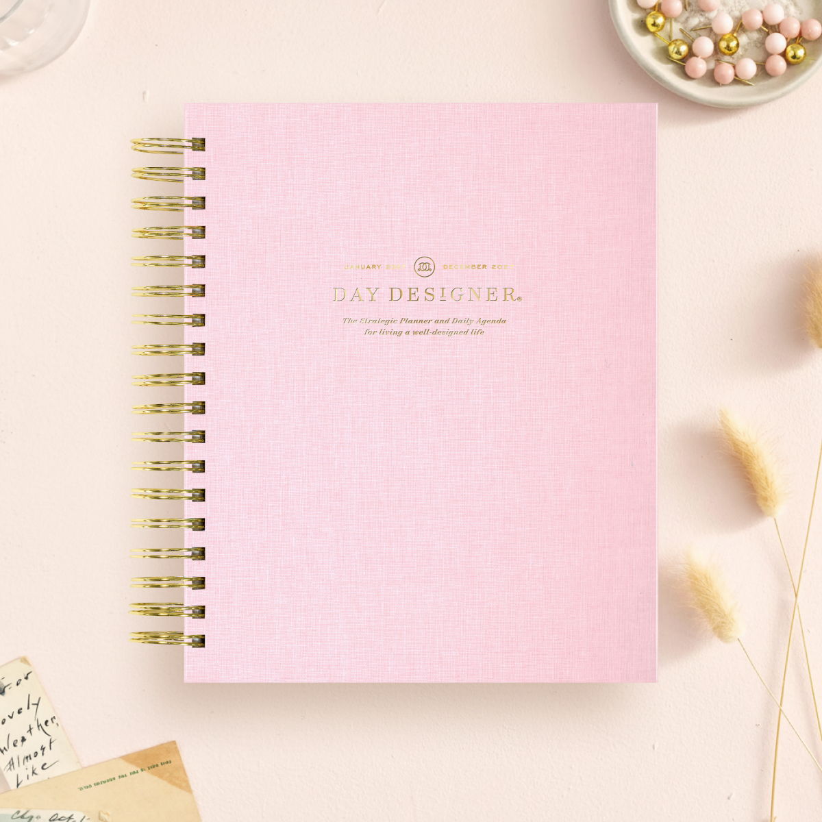 2027 day designer planner featuring a blush pink planner, gold binding on a beige background
